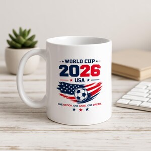 May include: A white ceramic mug featuring a graphic design with the text WORLD CUP 2026 USA and the phrase ONE NATION. ONE GAME. ONE DREAM. The design includes a soccer ball centered over a stylized American flag motif in red, white, and blue.