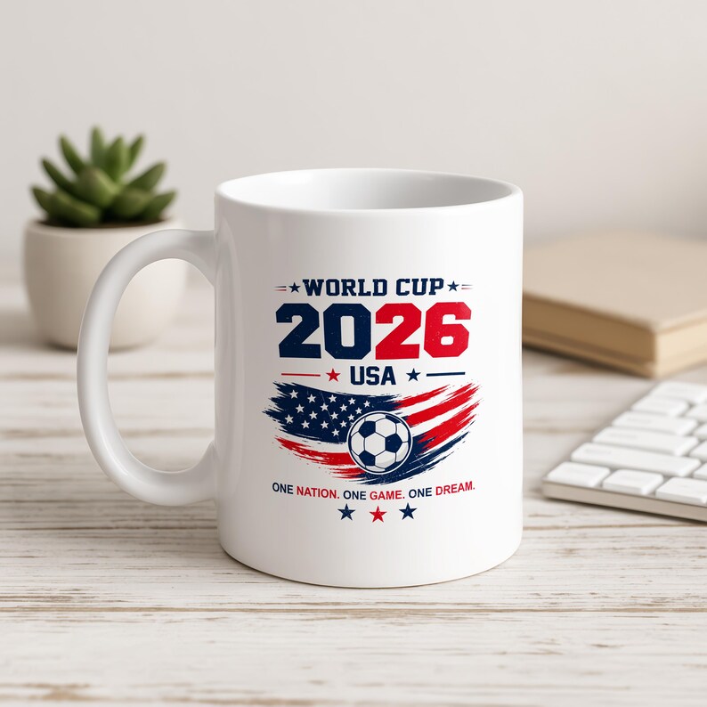 May include: A white ceramic mug featuring a graphic design with the text WORLD CUP 2026 USA and the phrase ONE NATION. ONE GAME. ONE DREAM. The design includes a soccer ball centered over a stylized American flag motif in red, white, and blue.
