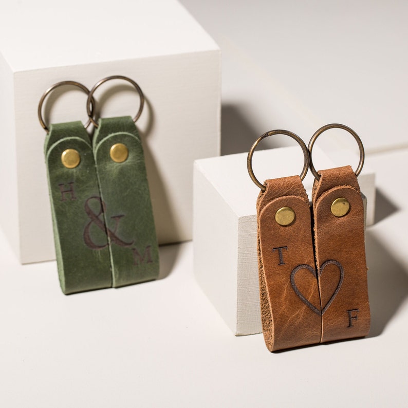 May include: Two leather keychains with metal rings. One keychain is green with the initials "H & M" engraved. The other keychain is brown with the initials "T & F" and a heart engraved.