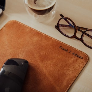 May include: A brown leather desk mat with a stitched edge and the name "Frank J. Adams" embossed in the lower right corner. A black computer mouse and a pair of tortoiseshell glasses are on the mat.