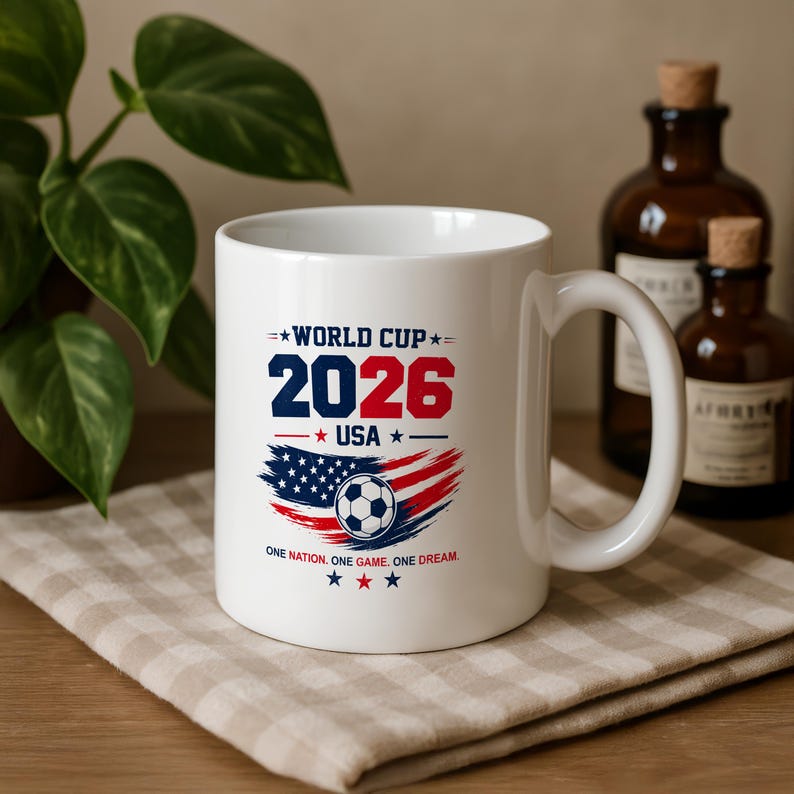May include: A white ceramic mug featuring a World Cup 2026 USA graphic. The design includes the text 'WORLD CUP 2026 USA', a soccer ball centered over a red, white, and blue brushstroke American flag motif, and the slogan 'ONE NATION. ONE GAME. ONE DREAM.'