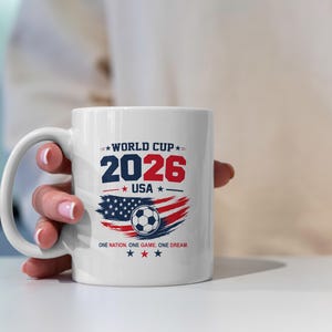 May include: A white ceramic mug featuring a World Cup 2026 USA graphic. The design includes the text WORLD CUP 2026 USA, a soccer ball, a stylized American flag, and the slogan ONE NATION. ONE GAME. ONE DREAM. with red, white, and blue stars.