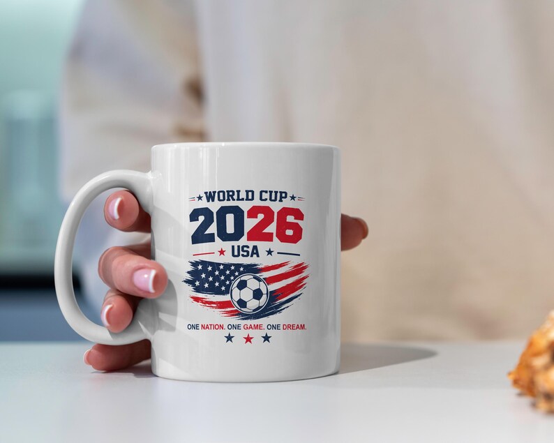 May include: A white ceramic mug featuring a World Cup 2026 USA graphic. The design includes the text WORLD CUP 2026 USA, a soccer ball, a stylized American flag, and the slogan ONE NATION. ONE GAME. ONE DREAM. with red, white, and blue stars.