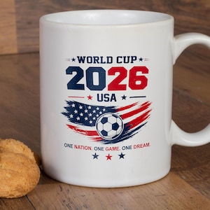 May include: A white ceramic mug featuring a World Cup 2026 USA graphic in blue and red. The design includes a soccer ball, a stylized American flag, and the text: WORLD CUP 2026 USA, and ONE NATION. ONE GAME. ONE DREAM. with star accents.