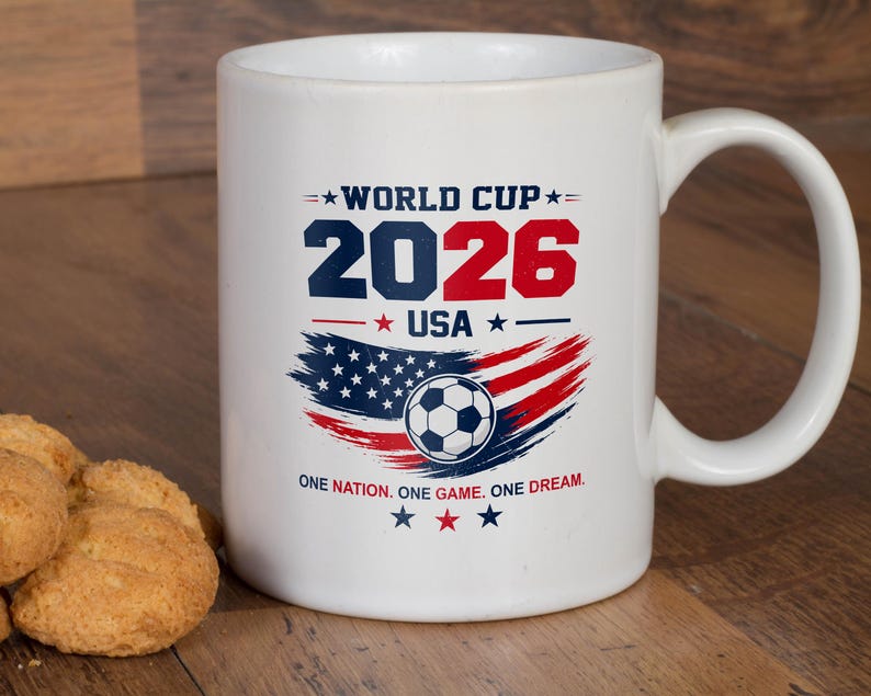 May include: A white ceramic mug featuring a World Cup 2026 USA graphic in blue and red. The design includes a soccer ball, a stylized American flag, and the text: WORLD CUP 2026 USA, and ONE NATION. ONE GAME. ONE DREAM. with star accents.