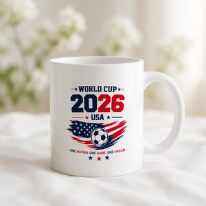 May include: A white ceramic mug featuring a World Cup 2026 USA graphic. The design includes the text WORLD CUP 2026 USA, a soccer ball, a stylized American flag in red, white, and blue, and the text ONE NATION. ONE GAME. ONE DREAM.