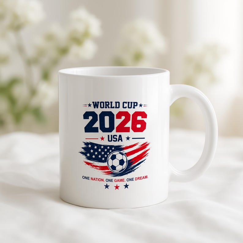 May include: A white ceramic mug featuring a World Cup 2026 USA graphic. The design includes the text WORLD CUP 2026 USA, a soccer ball, a stylized American flag in red, white, and blue, and the text ONE NATION. ONE GAME. ONE DREAM.