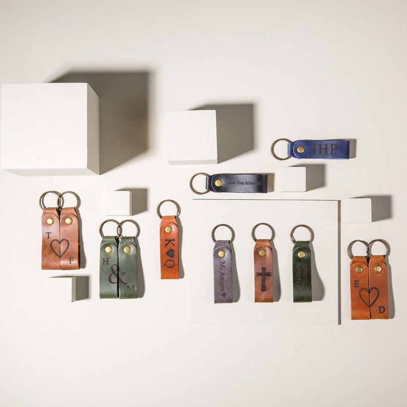 May include: Assortment of leather keychains with various engraved designs, including hearts, initials, a cross, and a playing card. The keychains are displayed on a white surface.