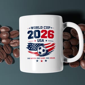 May include: A white ceramic mug featuring the text WORLD CUP 2026 USA in blue and red, a graphic of a soccer ball over a stylized American flag, and the text ONE NATION. ONE GAME. ONE DREAM. with small blue and red stars above and below the design.
