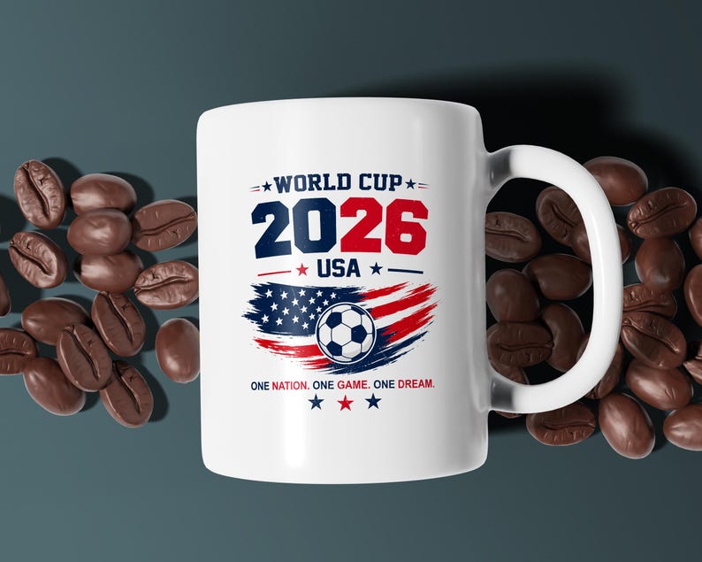 May include: A white ceramic mug featuring the text WORLD CUP 2026 USA in blue and red, a graphic of a soccer ball over a stylized American flag, and the text ONE NATION. ONE GAME. ONE DREAM. with small blue and red stars above and below the design.