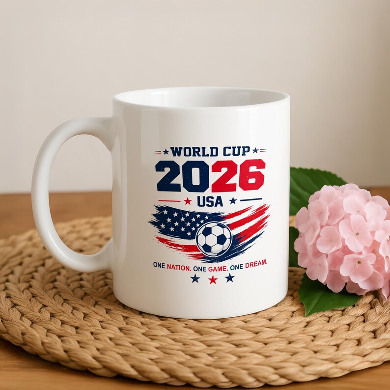May include: A white ceramic mug featuring a graphic with the text WORLD CUP 2026 USA and ONE NATION. ONE GAME. ONE DREAM. The design includes a soccer ball centered over a stylized American flag in red, white, and blue with star accents.
