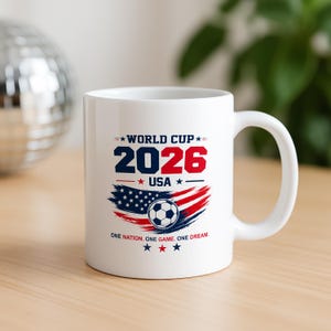 May include: A white ceramic mug featuring a World Cup 2026 USA graphic. The design includes the text 'WORLD CUP 2026 USA', a soccer ball, a stylized American flag in red, white, and blue, and the phrase 'ONE NATION. ONE GAME. ONE DREAM.'