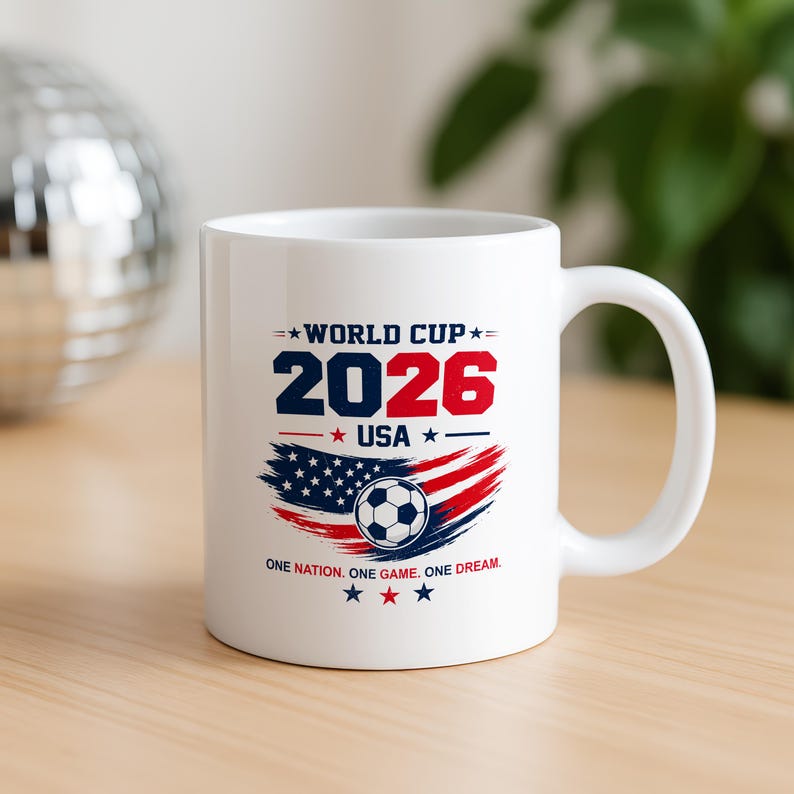 May include: A white ceramic mug featuring a World Cup 2026 USA graphic. The design includes the text 'WORLD CUP 2026 USA', a soccer ball, a stylized American flag in red, white, and blue, and the phrase 'ONE NATION. ONE GAME. ONE DREAM.'