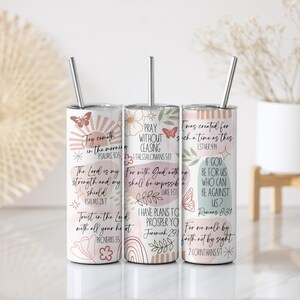 Puede incluir: Tres vasos aislados de acero inoxidable con tapas y pajitas. Cada vaso presenta un dise&ntilde;o &uacute;nico con citas inspiradoras, ilustraciones florales y de mariposas, y referencias b&iacute;blicas. Los vasos son blancos con detalles en rosa y beige.