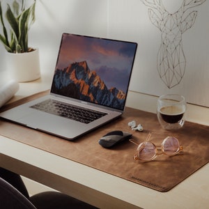 May include: A brown leather desk mat with the text "Remarkable Leather" printed on it. A laptop computer, a wireless mouse, a pair of glasses, a glass of coffee, and a pair of white earbuds are on the desk mat.