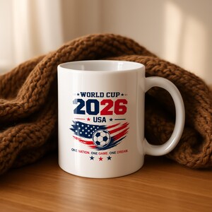 May include: A white ceramic mug featuring the text WORLD CUP 2026 USA in blue and red. Below the text is a graphic of a soccer ball over a stylized American flag design. The bottom text reads ONE NATION. ONE GAME. ONE DREAM. with three small stars.