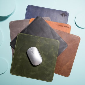 May include: A set of six leather mouse pads in various colors, including green, brown, gray, black, and blue. The mouse pads are stacked on top of each other, with a wireless mouse on top of the green one. The mouse pads have a stitched edge and are personalized with the text "Frank J. Adams" and a monogram.
