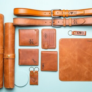 May include: A set of brown leather goods including a wallet, keychains, belts, and a desk mat. The items are personalized with initials and a heart symbol. The desk mat is also personalized with the name "Frank J Adams".