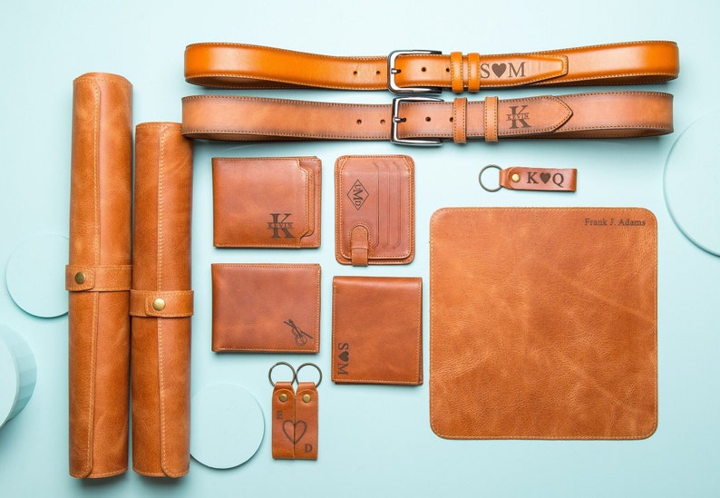 May include: A set of brown leather goods including a wallet, keychains, belts, and a desk mat. The items are personalized with initials and a heart symbol. The desk mat is also personalized with the name "Frank J Adams".