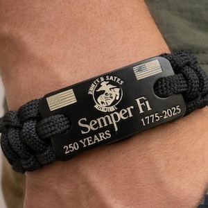 Può includere: Braccialetto in paracord nero con una placca metallica con le scritte "Semper Fi" e "250 YEARS 1775-2025". Il braccialetto è indossato su un polso, con un motivo mimetico visibile sullo sfondo.