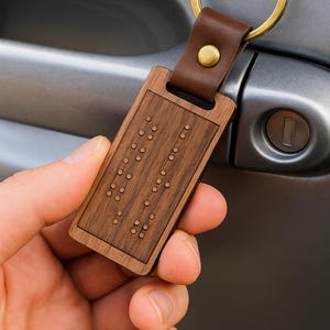 May include: A rectangular wooden keychain with braille dots. The keychain has a brown leather strap attached to a gold-colored ring. The wooden portion is a medium brown color.