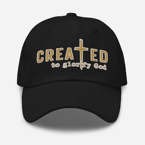 Könnte beinhalten: Schwarze Baseballkappe mit dem gestickten Text "CREATED to glorify God" in goldfarbenem Garn. Das Wort "CREATED" ist über einem goldenen Kreuz gewölbt, der Rest des Textes darunter. Die Kappe hat einen gebogenen Schirm und einen Knopf oben.