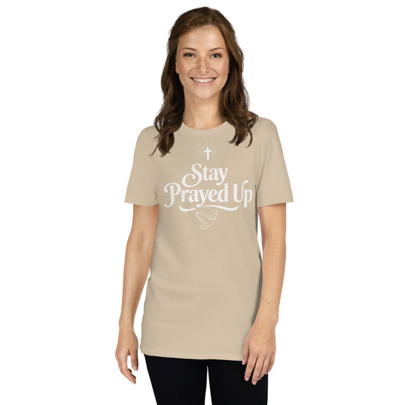 May include: A beige t-shirt with the text "Stay Prayed Up" in white script, featuring a cross and a dove. The shirt is short-sleeved and worn by a person. The overall style is casual and faith-based.