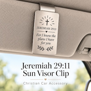 May include: A rectangular silver-toned metal sun visor clip featuring an engraved cross, the text Jeremiah 29:11, and the phrase For I know the plans I have for you. The clip is attached to a beige car sun visor with a smooth, brushed finish.