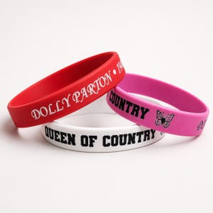 May include: Three silicone wristbands in red, pink, and white. The red band features white text reading DOLLY PARTON. The pink band has black text reading COUNTRY and a butterfly graphic. The white band features black text reading QUEEN OF COUNTRY.