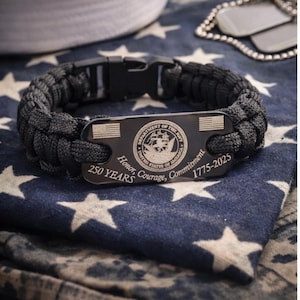 May include: A black paracord bracelet with a black metal plate. The plate is engraved with the words "DEPARTMENT OF THE NAVY UNITED STATES OF AMERICA" and "Honor, Courage, Commitment 1775-2025". The bracelet is on a blue and white star-patterned fabric.