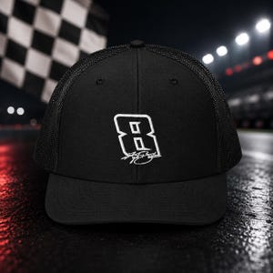 Pode incluir: Boné de caminhoneiro preto com o número 8 e assinatura bordados em branco. O boné apresenta uma parte traseira em malha e uma aba curva. O fundo inclui uma bandeira quadriculada e luzes de pista.