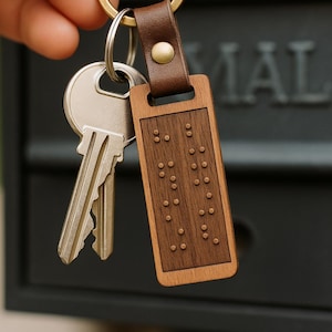 May include: A wooden rectangular keychain with a brown leather strap and brass hardware. The wooden portion features a braille pattern. Two keys are attached to the keychain.