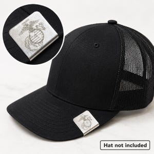 May include: A silver-toned metal emblem featuring an engraved United States Marine Corps Eagle, Globe, and Anchor insignia. The rectangular metal piece is designed to clip onto the brim of a black mesh-back baseball cap, shown in the image.