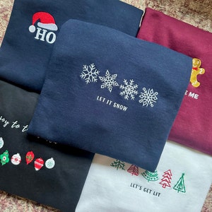 May include: Five different Christmas themed sweatshirts. The first is navy blue with a red Santa hat and the text "HO". The second is navy blue with three snowflakes and the text "LET IT SNOW". The third is maroon with a gingerbread man and the text "ME". The fourth is black with a row of Christmas ornaments. The fifth is white with a row of green Christmas trees and the text "LET'S GET LIT".