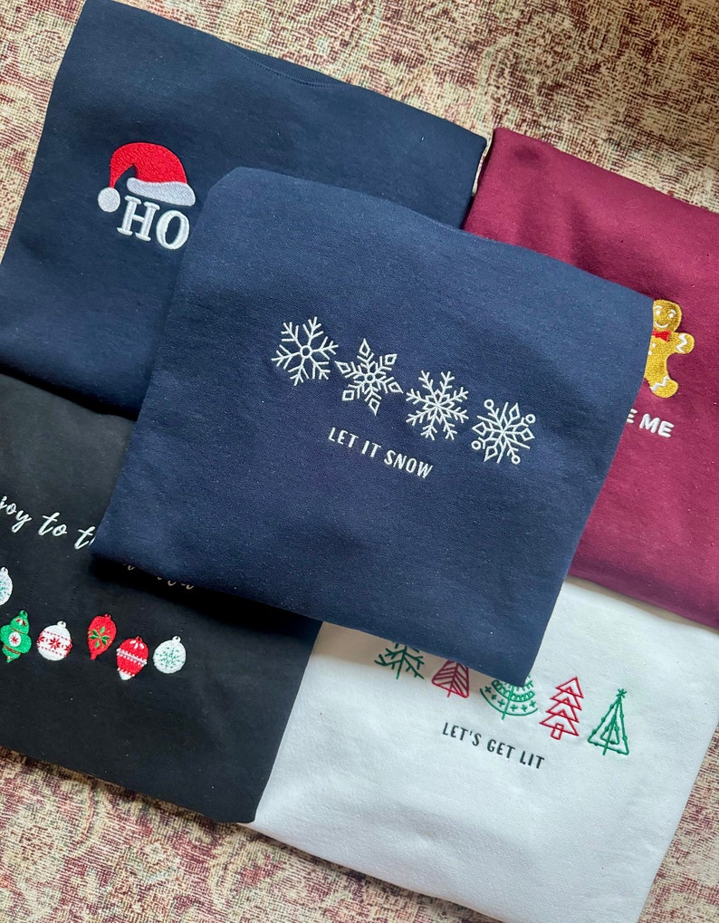 May include: Five different Christmas themed sweatshirts. The first is navy blue with a red Santa hat and the text "HO". The second is navy blue with three snowflakes and the text "LET IT SNOW". The third is maroon with a gingerbread man and the text "ME". The fourth is black with a row of Christmas ornaments. The fifth is white with a row of green Christmas trees and the text "LET'S GET LIT".