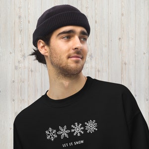 May include: A black sweatshirt with a white snowflake design and the text "LET IT SNOW". The model is wearing a black beanie.