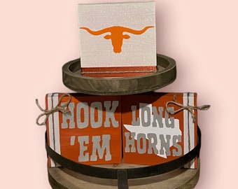 May include: A two-tiered wooden tray with a white and orange block on the top tier. The block has a silhouette of a longhorn steer. The bottom tier has two orange blocks with the words "Hook 'em Horns" in white lettering.