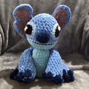 May include: A handmade blue crochet plush toy, resembling a cartoon character. The toy features large ears with brown accents, black eyes, and a smiling mouth. The plush is made of soft, textured yarn and is sitting on a gray surface.