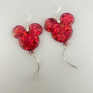May include: A pair of dangle earrings featuring red resin charms shaped like mouse heads with balloon-like silhouettes. The charms are filled with chunky red glitter and attached to silver-toned metal hooks with thin, curved silver-toned wire tails.