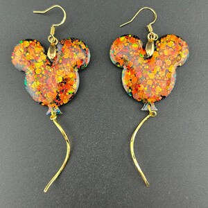 May include: A pair of dangle earrings featuring a mouse-shaped balloon design filled with orange, red, and gold glitter. Each earring hangs from a gold-toned hook and includes a thin, curved gold-toned wire extending from the bottom of the balloon.
