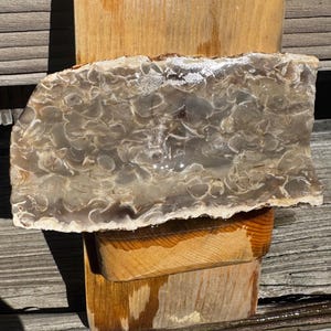 May include: A polished, translucent stone slab with a unique pattern of swirling, light gray and brown colors. The stone is displayed on a stack of wooden blocks, showcasing its natural texture and beauty. The stone's edges are rough, adding to its organic appeal.