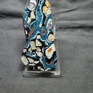 May include: A polished, irregularly shaped stone with a vibrant pattern of blue, black, white, and orange swirls. The stone is displayed on a clear acrylic stand, showcasing its unique, natural design. The stone's surface has a mix of textures and colors.