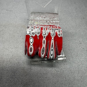 May include: A rectangular, decorative object with a red base and a patterned design of black, white, and gray circles and lines. The top section has a textured, silver and gray appearance. The object is displayed on a clear acrylic stand.