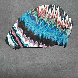 May include: A uniquely shaped, abstract art piece with a black base and vibrant, wavy patterns. The design incorporates shades of blue, green, red, white, and purple, creating a dynamic and colorful composition. The artwork is set against a gray backdrop.