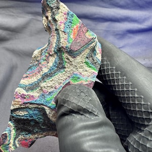 May include: A colorful, layered rock fragment with a variety of colors including blue, green, pink, and white. The rock is held by a gloved hand. The rock's surface has a textured appearance, with visible layers and patterns.