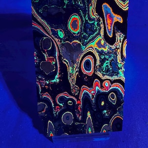 May include: A dark, rectangular stone slab with a unique, colorful pattern. The surface features swirling lines and circular shapes in shades of black, red, orange, green, and blue. The stone is set against a blue background.