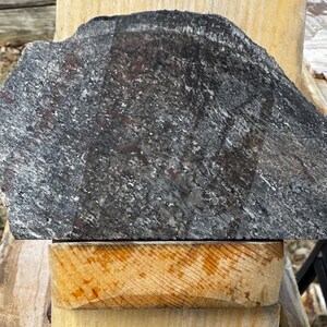 Puede incluir: Una gran pieza de piedra gris oscuro de forma irregular con una superficie texturizada. La piedra tiene vetas blancas y rojizas. Descansa sobre un bloque de madera de color claro, lo que sugiere una estética natural o rústica.