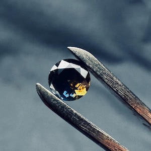 Puede incluir: Una gema facetada sostenida por unas pinzas metálicas. La gema muestra una mezcla de colores, incluyendo amarillo, azul y negro, con un corte brillante. El fondo es gris liso y apagado.