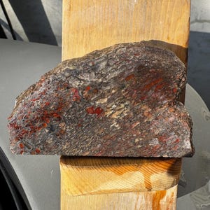 Puede incluir: Una roca grande, de forma irregular, con una superficie gris oscura y marrón, acentuada con vetas y manchas rojas. La roca descansa sobre un bloque de madera de color claro, mostrando sus texturas y colores naturales. Es un espécimen geológico único.