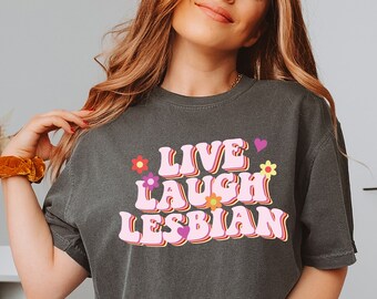 Puede incluir: Una camiseta gris oscuro con el texto "Live Laugh Lesbian" en fuente retro rosa con flores y corazones coloridos.